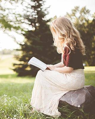 A young woman finds solace in a book, seated on a smooth polished rock amidst lush grass, with a tranquil evergreen backdrop.